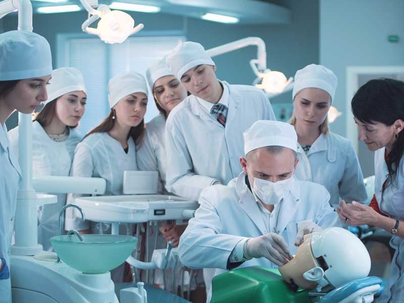 Estudiantes para cirujano dentista observan atentos a su profesor haicendo una práctica en un simulador