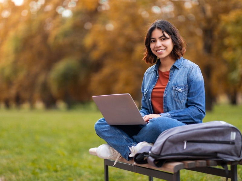 Estudiante con laptop estudiando en línea al aire libre en un parque durante el otoño.