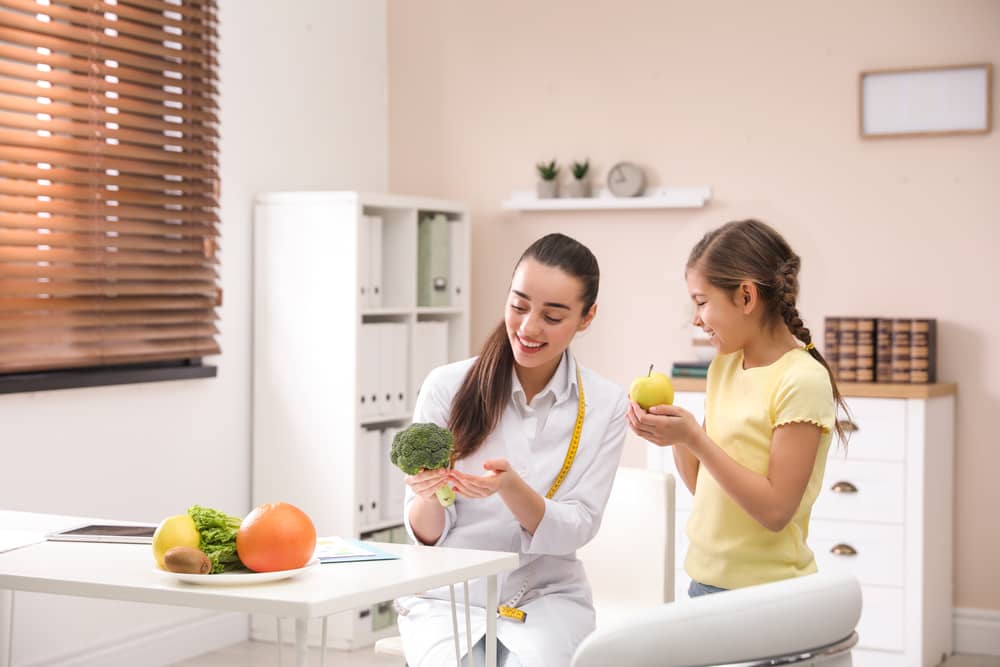 Nutrióloga enseñando a niña sobre alimentación saludable con frutas y verduras en consultorio.