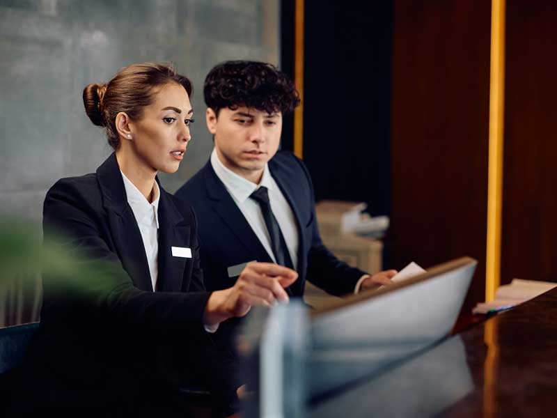 Conserje femenina asistiendo a la recepcionista en el mostrador del hotel, ofreciendo un servicio personalizado y atención al cliente en un entorno de hospitalidad.