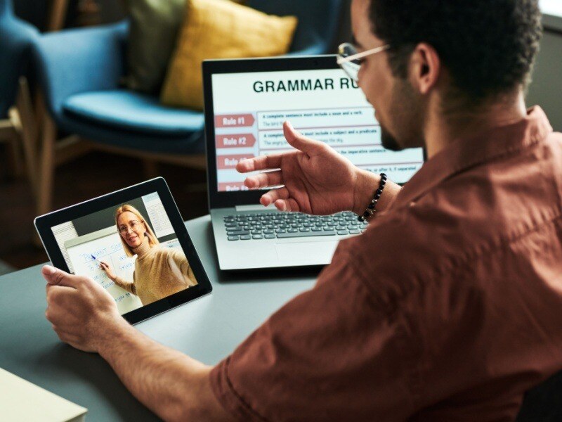 Hombre tomando una clase de inglés online en su tablet, con gramática en la pantalla de su computadora, representando aprendizaje flexible y profesional.