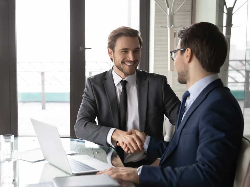 Dos profesionales en trajes de negocios estrechando manos en una oficina, representando el networking y colaboraciones en el ámbito empresarial