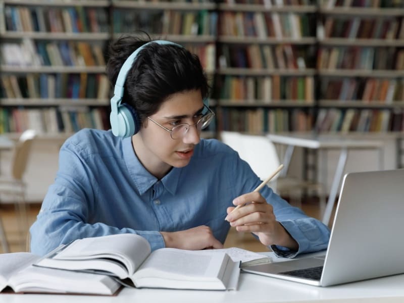 Joven estudiando en biblioteca con audífonos y laptop para el examen CENEVAL de preparatoria