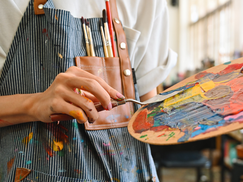 Persona mezclando colores en paleta de pintura con espátula y pinceles en delantal artístico