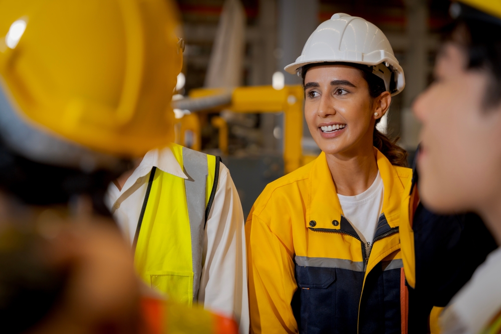 Ingeniera con casco blanco y uniforme de seguridad conversa con su equipo en una planta industrial.