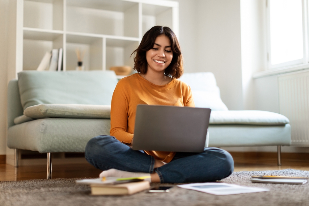Mujer utiliza una laptop sentada en la alfombra de su sala, rodeada de libros, apuntes y un cuaderno.